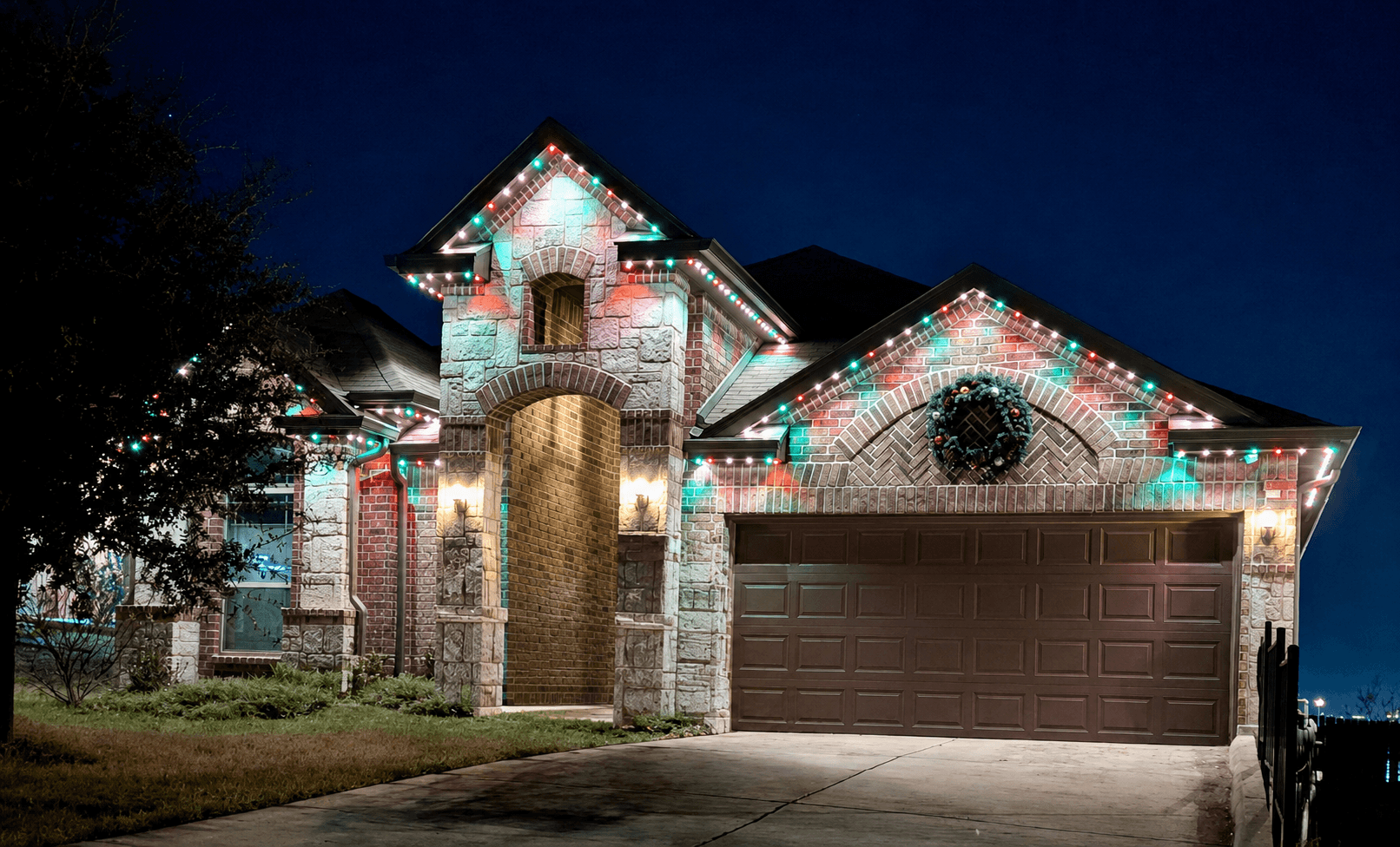 Customer-supplied LIT365 installation: a stone home's gables and garage outlined in multicolor permanent roofline lighting at night