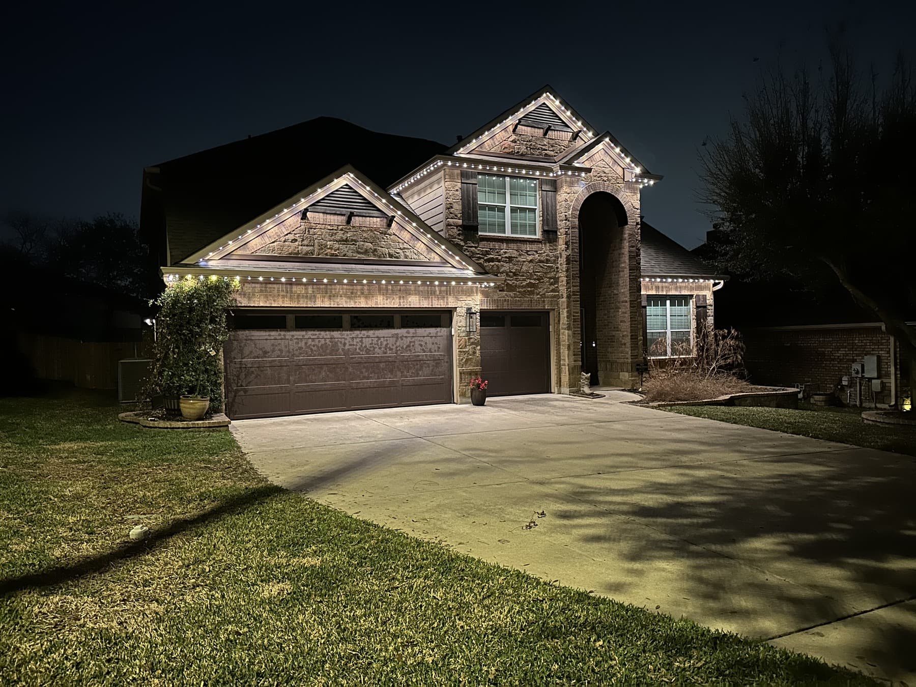 Customer-supplied LIT365 installation: a stone home's roof peaks, facade, and driveway under warm permanent roofline lighting at night